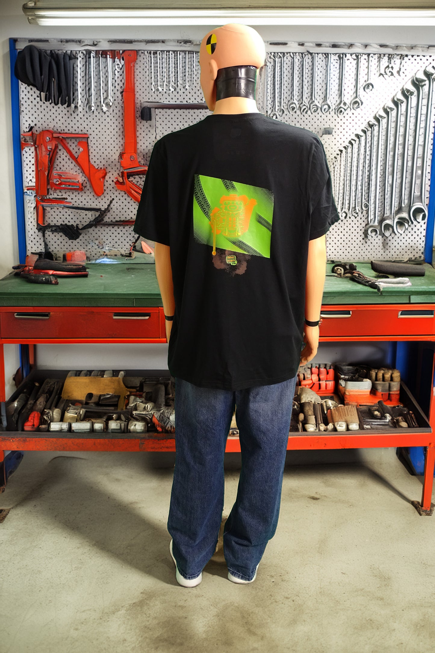  Crash test dummy mannequin in a workshop setting wearing a black t-shirt with the under side of a JDM engine with exhaust coming out and a small GKS logo in the middle of the smoke on the back.