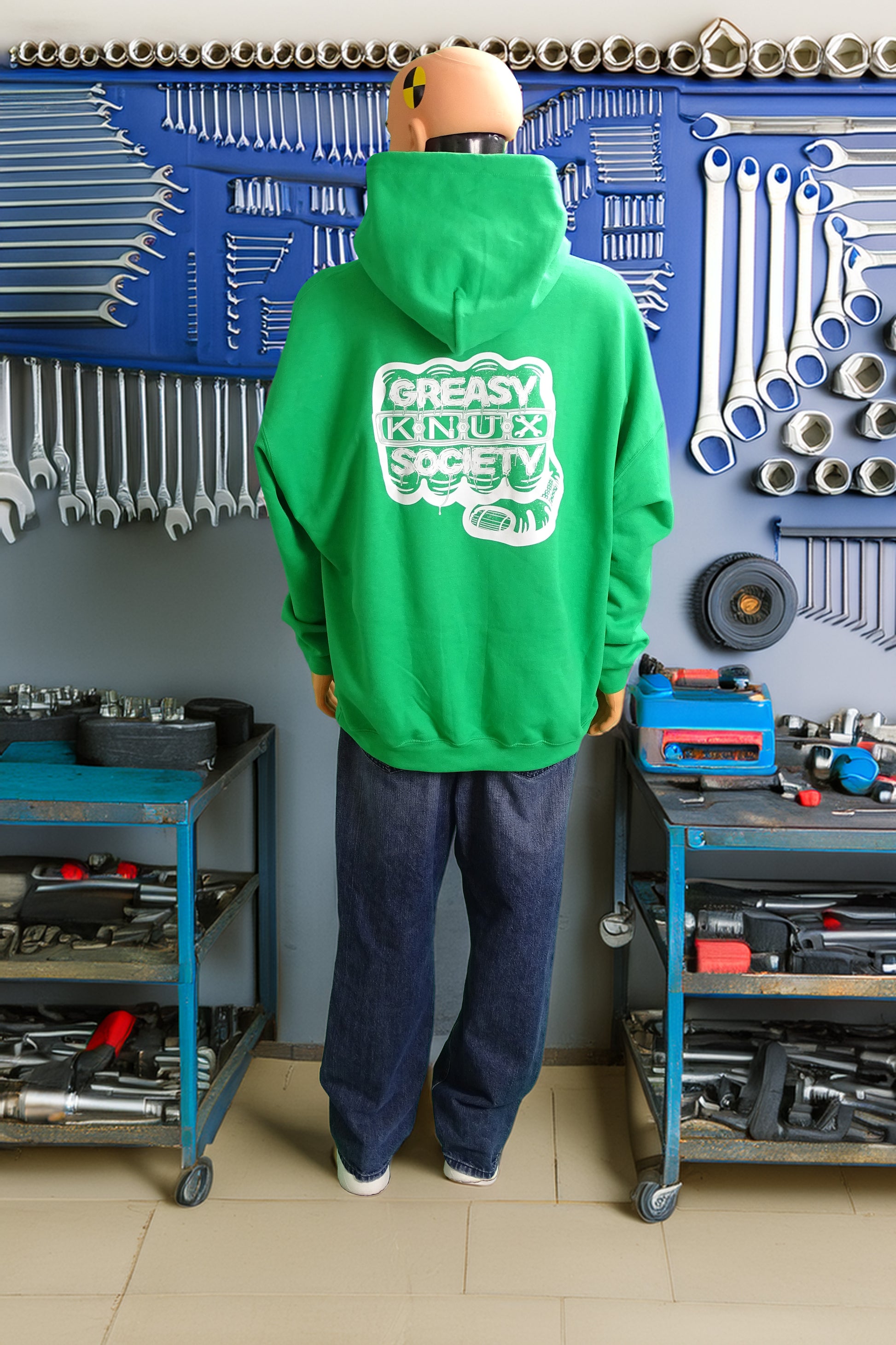 Crash test dummy mannequin in a workshop setting wearing a green hoodie with with a large white logo consisting of big fist with brass knuckles and bandage on the thumb with type reading Greasy Knux Society. Greasy and Society are stylized to look like dripping grease and Knux is made of various tools.