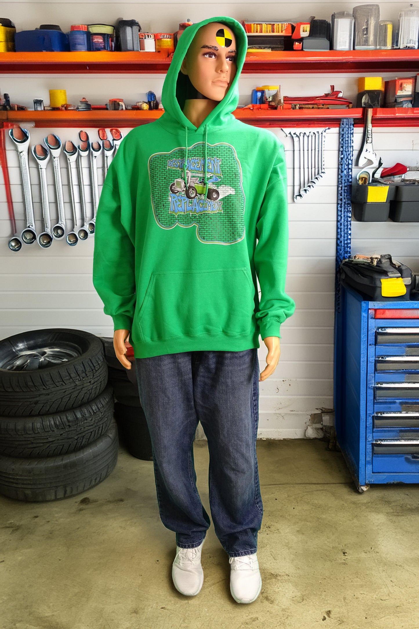 Crash test dummy mannequin in a workshop setting wearing a green hoodie with green and blue illustration of a retro coupe with a small engine and a giant turbo charger spinning it's wheels in front of a brick wall with a crash test dummy driving and graffiti styled text that reads Displacement Replacement.
