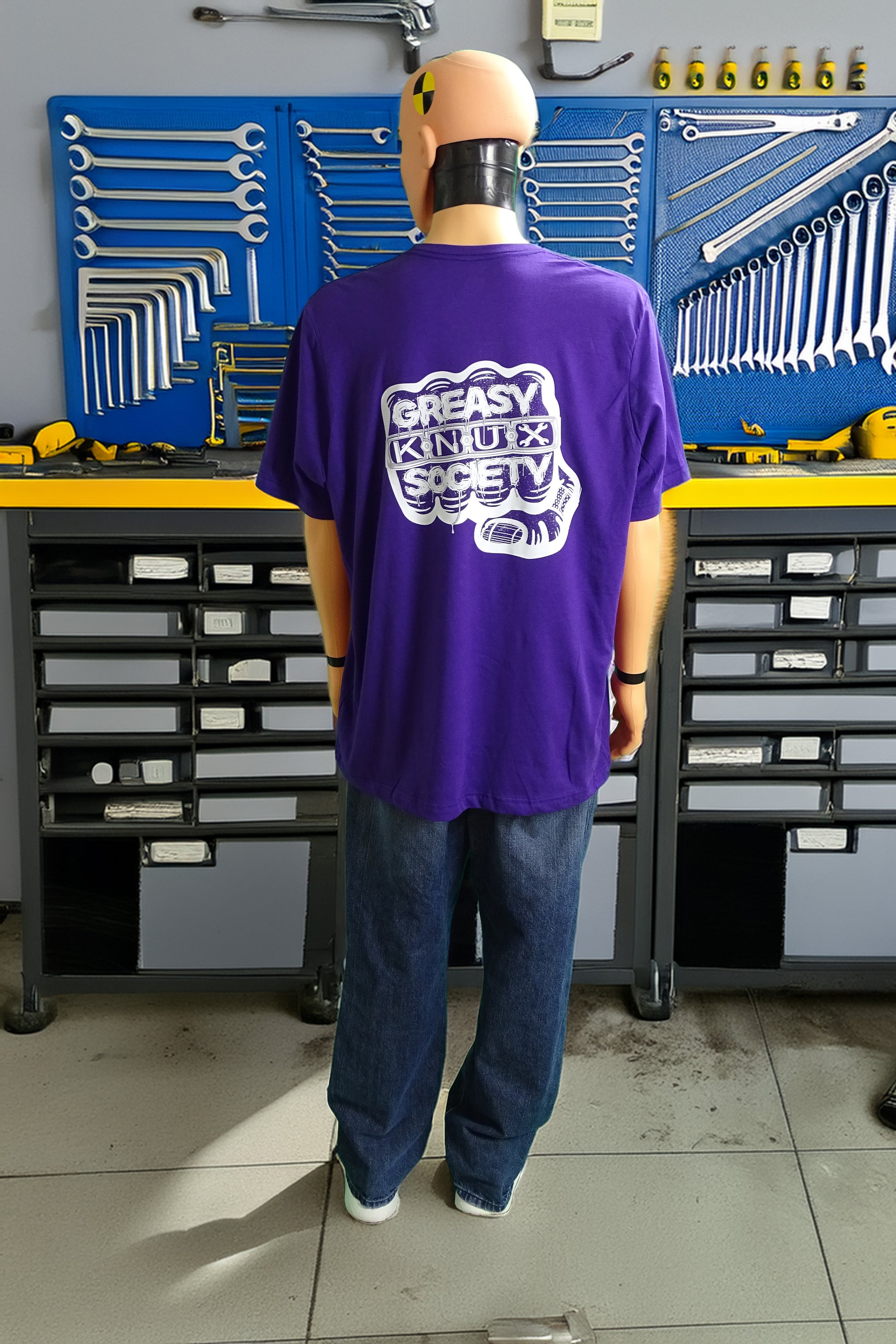 Crash test dummy mannequin in a workshop setting wearing a purple t-shirt with with a large white logo consisting of big fist with brass knuckles and bandage on the thumb with type reading Greasy Knux Society. Greasy and Society are stylized to look like dripping grease and Knux is made of various tools.